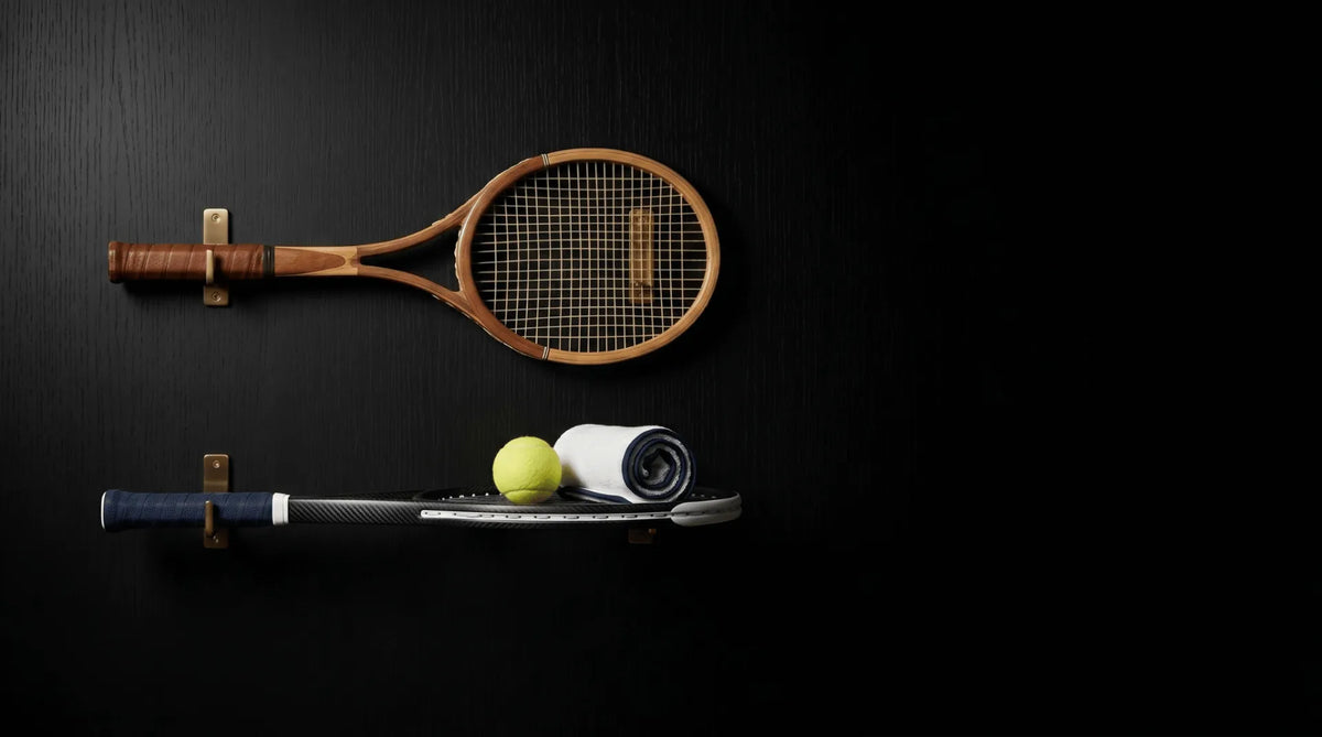 Two tennis rackets on a black wall, one wooden, one modern, with a tennis ball and towel